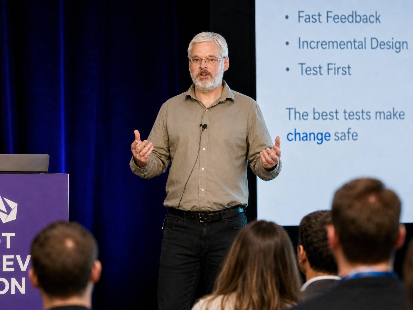 Dave Rooney speaking to an audience at a conference, gesturing beside a slide reading 'The best tests make change safe'