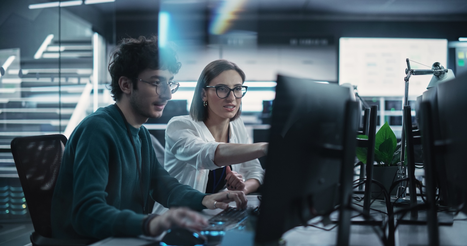 A coach leaning in beside a developer at his keyboard, pointing something out on his screen